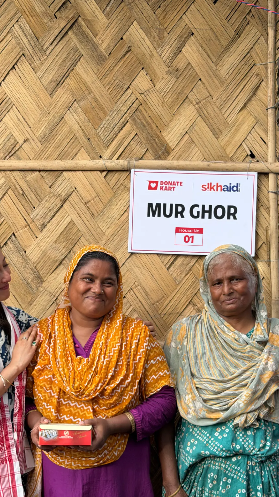 Two women from Assam stand with a Donatekart team member outside a rebuilt bamboo home marked Mur Ghor, House No. 01